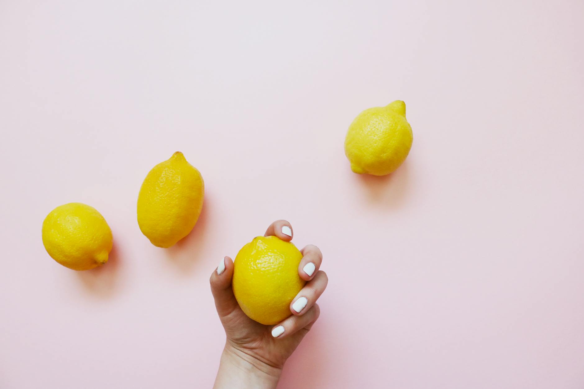 Una mano che tiene un limone fresco su sfondo rosa morbido, circondata da altri limoni.