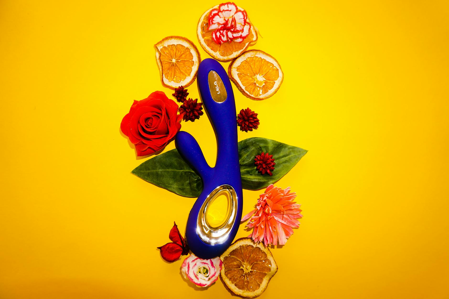 Vibratore blu circondato da fiori e fette di arancia su sfondo giallo