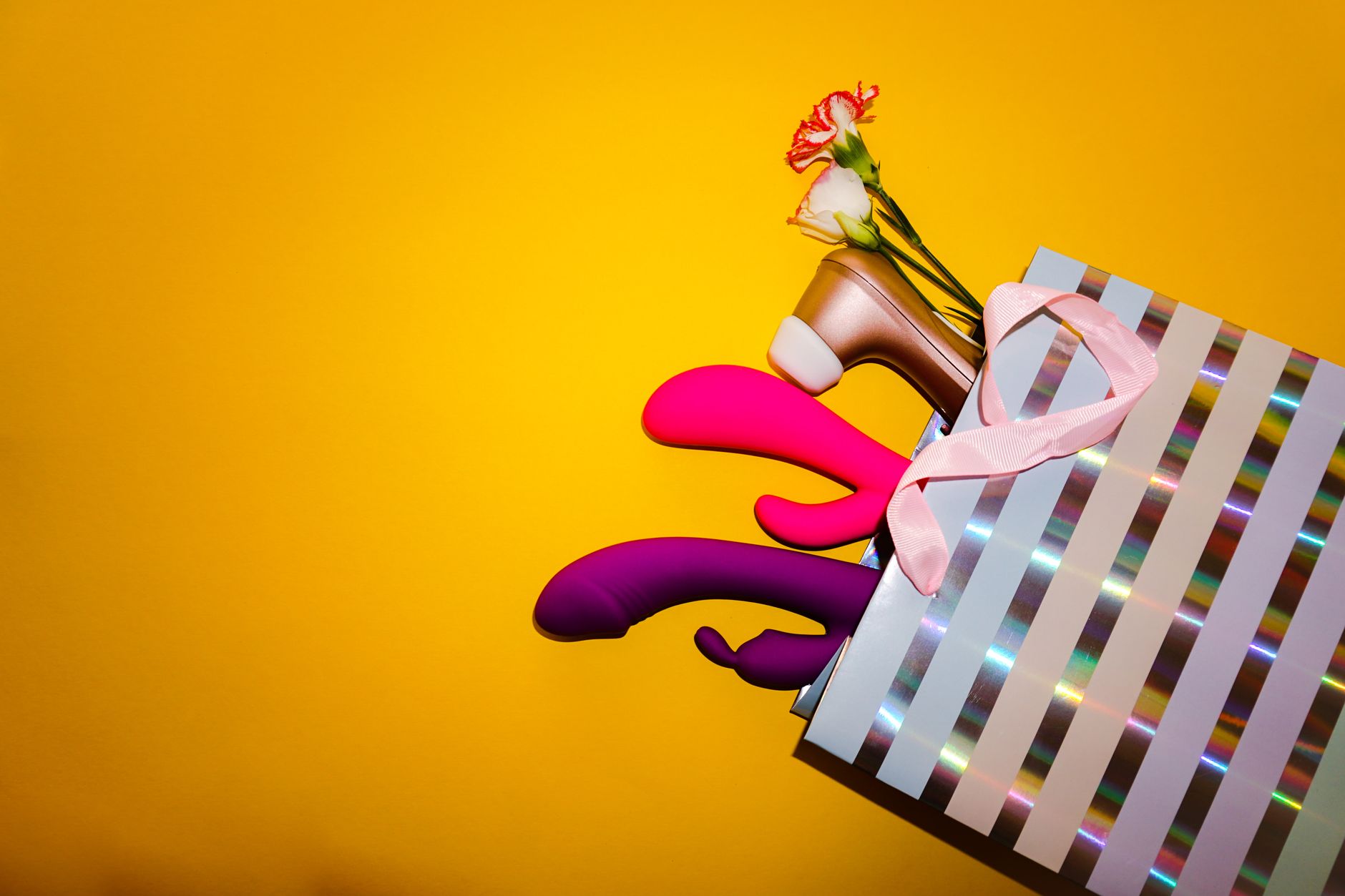 Vibratori colorati con fiori in una borsa regalo olografica su sfondo giallo vivace